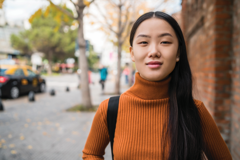 teenage female in orange turtleneck standing on a sidewalk on a sunny day